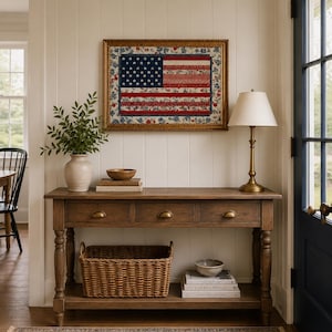 Pode incluir: Uma colcha com a bandeira americana emoldurada com uma moldura dourada est&aacute; pendurada acima de uma mesa de console de madeira. A mesa tem tr&ecirc;s gavetas, uma prateleira inferior com uma cesta de vime e uma l&acirc;mpada de lat&atilde;o. Um tapete estampado e um vaso com vegeta&ccedil;&atilde;o completam a cena.