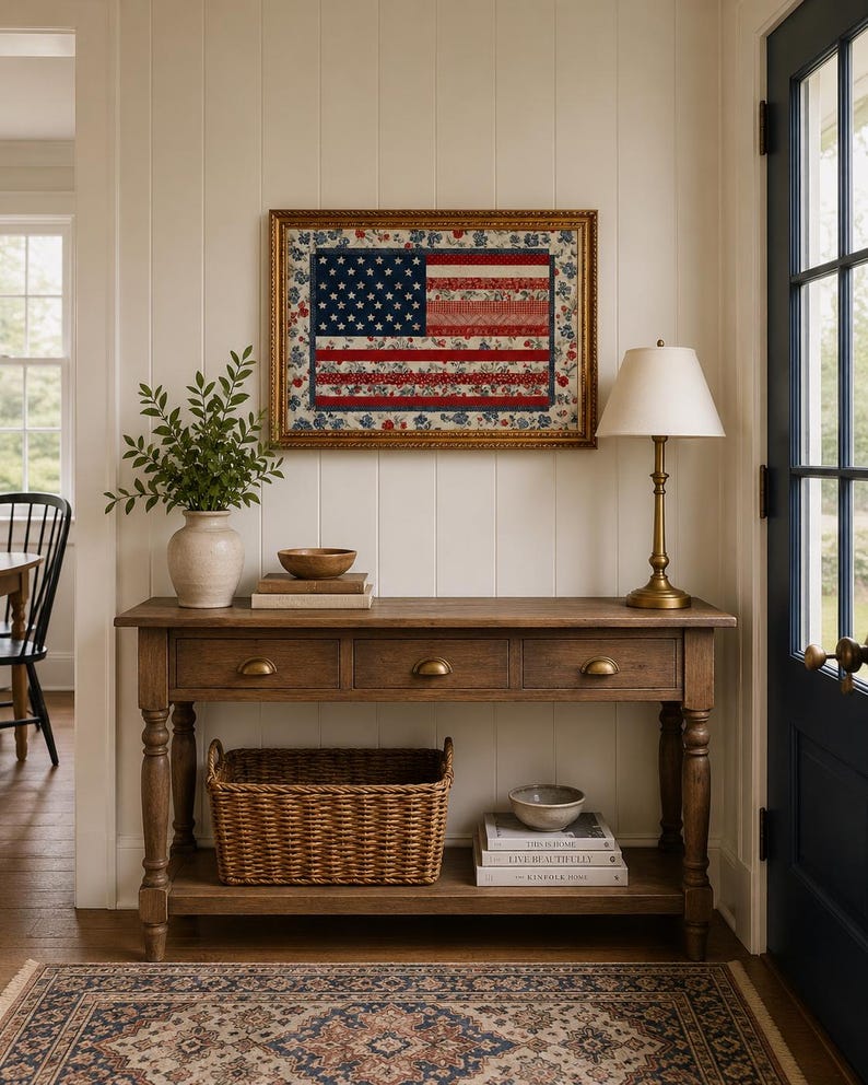 Pode incluir: Uma colcha com a bandeira americana emoldurada com uma moldura dourada est&aacute; pendurada acima de uma mesa de console de madeira. A mesa tem tr&ecirc;s gavetas, uma prateleira inferior com uma cesta de vime e uma l&acirc;mpada de lat&atilde;o. Um tapete estampado e um vaso com vegeta&ccedil;&atilde;o completam a cena.