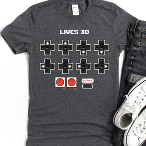 Peut inclure: Un t-shirt gris foncé avec un design de manette de jeu vidéo rétro. Le t-shirt présente le texte "LIVES 30" et un graphique d'une manette avec des boutons et un bouton de démarrage.