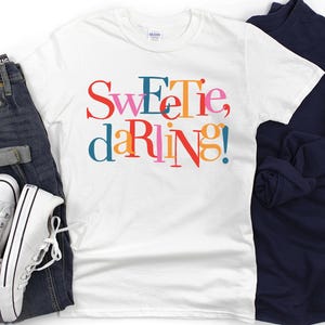 Puede incluir: Camiseta blanca con el texto "Sweet, darling!" en letras de colores. La camiseta está doblada y se muestra sobre un fondo blanco.