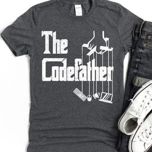Op de afbeelding: Donkergrijs t-shirt met de witte tekst "The Codefather". Het ontwerp toont de handen van een poppenspeler die een toetsenbord, muis en laptop bestuurt. Een paar witte sneakers en een jeans zijn ook zichtbaar.