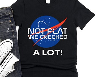 Puede incluir: Camiseta negra con un diseño gráfico azul y blanco inspirado en la NASA que dice "NOT FLAT WE CHECKED A LOT!". El diseño incluye un círculo azul con estrellas blancas, un arco blanco y una línea roja. La camiseta se muestra con vaqueros azules y zapatillas blancas.