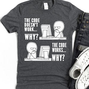 May include: A dark grey t-shirt featuring a humorous graphic. The white text reads "THE CODE DOESN'T WORK... WHY?" and "THE CODE WORKS... WHY?" with a meme-style illustration of a person at a computer. Includes jeans and white trainers.