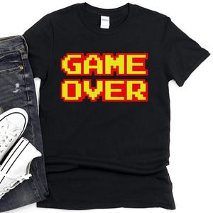 Op de afbeelding: Zwart t-shirt met de woorden "GAME OVER" in gepixelde gele en rode letters. Het shirt wordt getoond met een blauwe jeans en witte sneakers.
