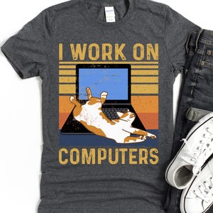 Op de afbeelding: Donkergrijs t-shirt met de tekst "I WORK ON COMPUTERS". De afbeelding toont een laptop met een kat die erop ligt. De kat is oranje en wit, en de achtergrond heeft horizontale strepen in geel, oranje en blauw.