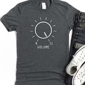 Könnte beinhalten: Dunkelgraues T-Shirt mit einem weißen Grafikdesign eines Lautstärkereglers. Das Design zeigt die Zahlen 0 und 11, mit dem Wort "VOLUME" darunter. Ein schlichtes, auffälliges Design.