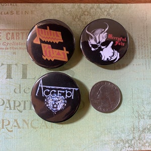 May include: Three black and white button pins with band logos. The pins feature the logos for Judas Priest, Accept, and Mercyful Fate. The pins are arranged in a triangular shape on a green and white patterned surface.