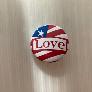 Pu&ograve; includere: Un bottone rotondo con un design a bandiera americana rosso, bianco e blu. La parola "Love" &egrave; stampata in bianco su uno striscione rosso sopra la bandiera.