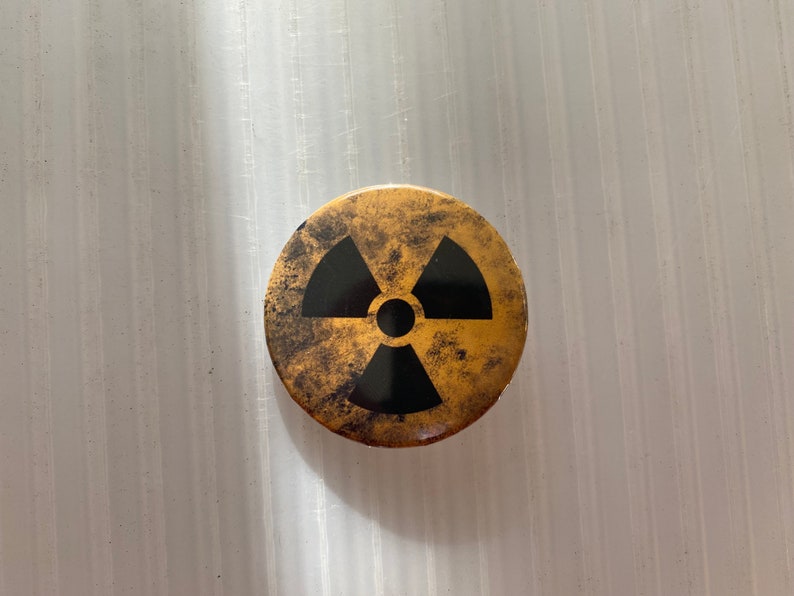 Pu&ograve; includere: Uno spillo rotondo color oro con un simbolo di radiazione nero. Lo spillo ha un aspetto usurato e invecchiato.