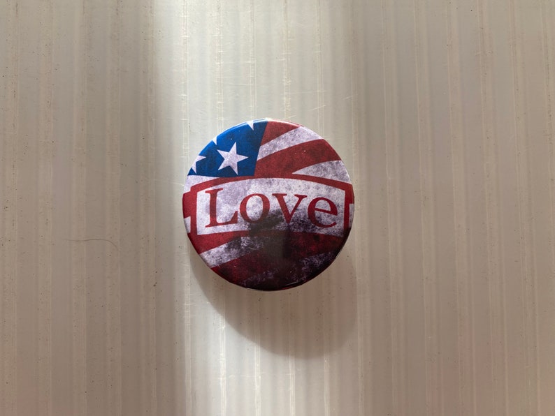 Pu&ograve; includere: Un bottone rotondo con un designo di bandiera americana usurato e la parola "Love" in lettere bianche.
