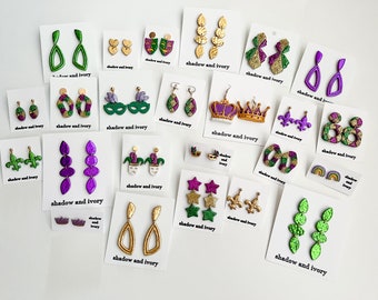 May include: A selection of handmade earrings in diverse styles, such as teardrops, crowns, and masks. The earrings are adorned with green, purple, gold, and glitter details. The cards display the text "shadow and ivory".