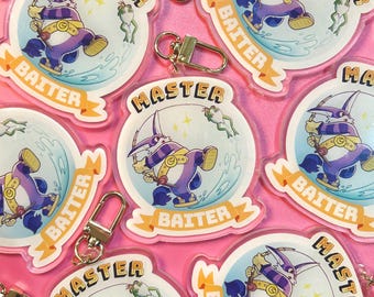 Op de afbeelding: Een verzameling heldere acryl sleutelhangers met een stripfiguur die aan het vissen is. Het personage is paars en wit, met de woorden "MASTER" en "BAITER" op de sleutelhanger. Elke sleutelhanger heeft een goudkleurige sluiting.