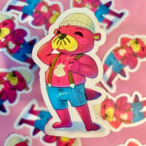 Peut inclure: Un autocollant représentant une loutre de dessin animé avec une fourrure rose, un museau jaune et un bonnet crème. La loutre porte un short en jean bleu, des bretelles marron et un pendentif coquillage. D'autres autocollants sont visibles en arrière-plan sur une surface rose.