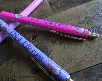 May include: Three pens with butterfly designs and the text "Best Life Ever." One pen is pink with glitter, another is purple, and the third is a lighter pink. The pens have gold-colored accents and are on a wooden surface.
