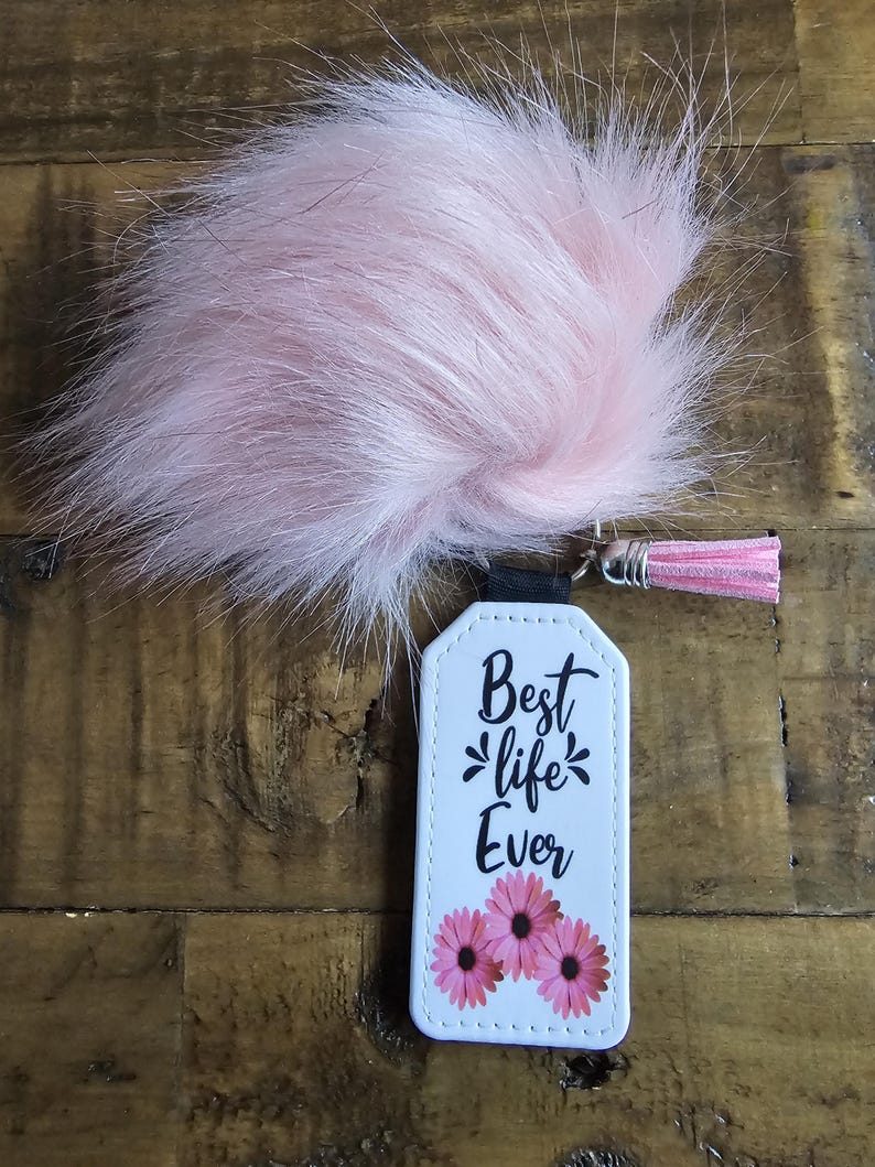 Pode incluir: Chaveiro de couro branco com um pompom rosa peludo. O chaveiro tem o texto "Best life Ever" e tr&ecirc;s flores rosas.