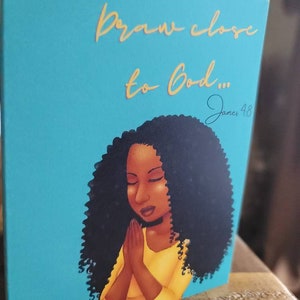 May include: A teal blue card with a gold inscription that reads "Draw close to God... James 4:8". The card features a digital illustration of a Black woman with long curly hair, praying with her hands clasped together.