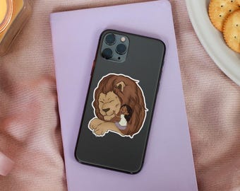 May include: A black smartphone with a sticker featuring a lion hugging a person. The sticker has a brown lion with a light brown mane and a person in a white shirt and purple skirt. The phone rests on a purple notebook.
