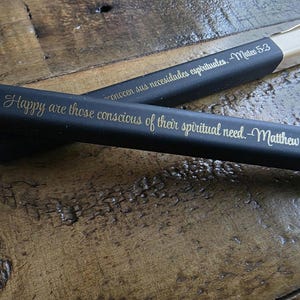 Puede incluir: Dos bolígrafos negros con letras doradas. Uno de los bolígrafos dice "Happy are those conscious of their spiritual need. -Matthew." El otro bolígrafo dice "conocen sus necesidades espirituales. -Mateo 5:3."