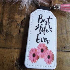 Pode incluir: Chaveiro de couro branco com o texto "Best life Ever" e tr&ecirc;s flores cor-de-rosa.