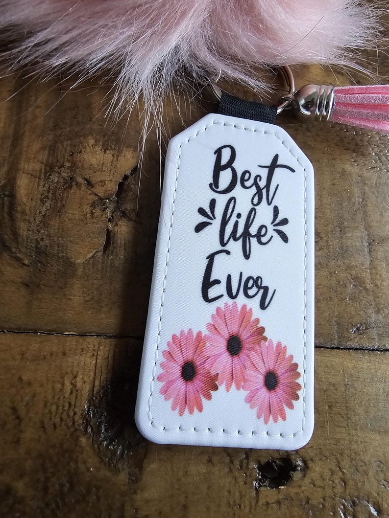 Pode incluir: Chaveiro de couro branco com o texto "Best life Ever" e tr&ecirc;s flores cor-de-rosa.