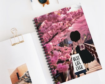 May include: Spiral-bound notebook featuring a vibrant illustration of a person walking on a bridge under pink cherry blossom trees. The notebook cover has the text "BEST LIFE EVER" on a small cart.