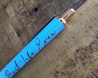 May include: A close-up of a pen with a light blue, glittery barrel. The pen has the words "Best Life Ever" in black script. The pen's tip and clip are gold-colored, and the background is a wooden surface.