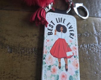 May include: A rectangular keychain with a woman illustration, the text "BEST LIFE EVER!", and a red fluffy pom-pom. The woman is wearing a white shirt, red skirt, and black heels. The background is a floral pattern.