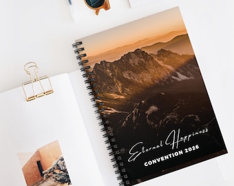 May include: A spiral-bound notebook with a mountain landscape cover. The cover features a sunset over a mountain range with the text "Eternal Happiness Convention 2026". The notebook is open, revealing blank pages and a gold paper clip.