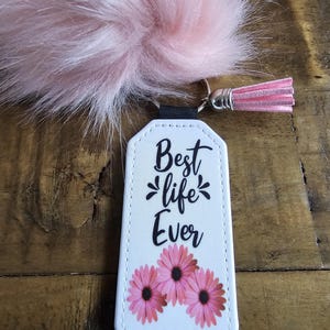 Pode incluir: Chaveiro de couro branco com um pompom peludo rosa. O chaveiro tem o texto "Best life Ever" e tr&ecirc;s flores rosas.