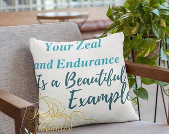 May include: White decorative pillow with teal text that reads "Your Zeal and Endurance Is a Beautiful Example." The pillow has a gold floral design in the lower left corner and is on a gray chair.