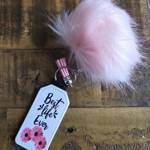 Pode incluir: Chaveiro branco com um pompom rosa peludo. O chaveiro tem uma etiqueta branca com texto preto que diz "Best life Ever" e flores rosas.
