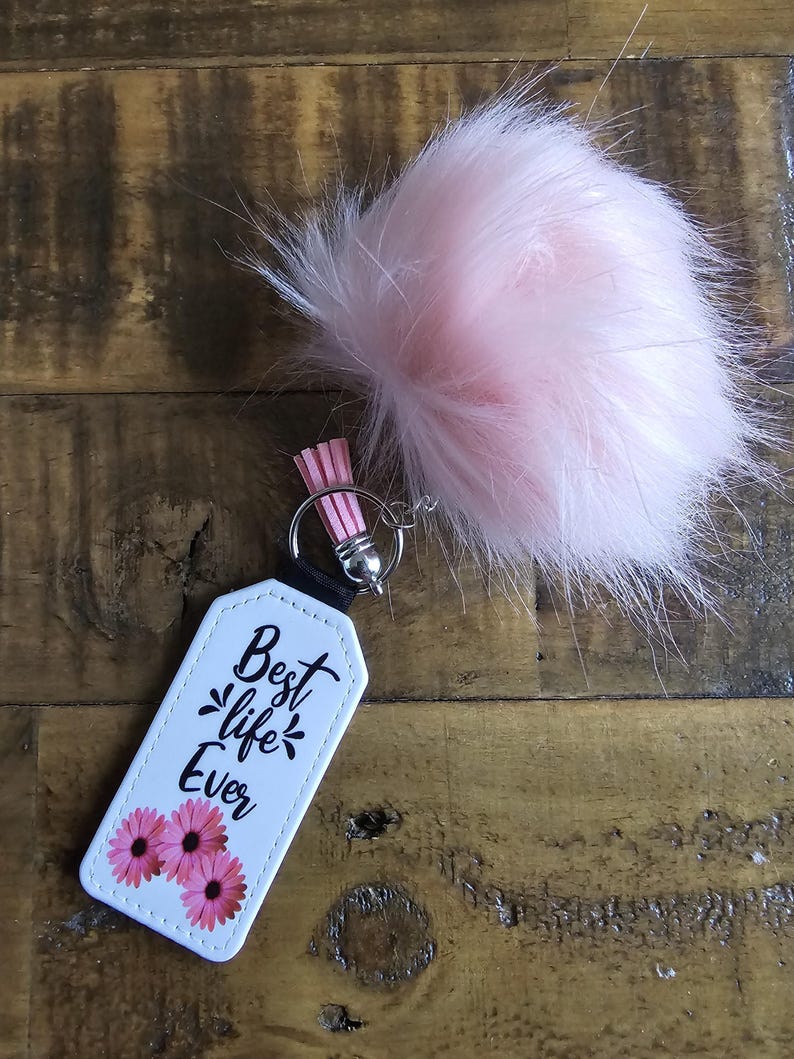 Pode incluir: Chaveiro branco com um pompom rosa peludo. O chaveiro tem uma etiqueta branca com texto preto que diz "Best life Ever" e flores rosas.