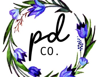 May include: A circular watercolor wreath composed of green leaves and vibrant blue bell-shaped flowers with small yellow accents at the bottom. Centered within the floral arrangement is the black text reading pd co. in a simple script and sans-serif font.