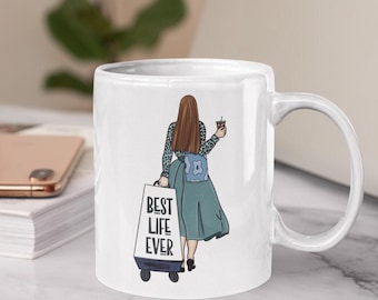 May include: White ceramic mug featuring an illustration of a person with long brown hair, a blue backpack, and a teal skirt, pulling a suitcase that reads "BEST LIFE EVER." The person is holding a coffee cup.
