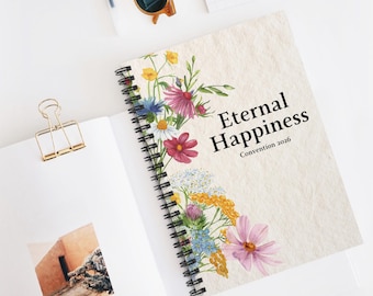 May include: A spiral-bound notebook with a floral design and the text "Eternal Happiness Convention 2026." The cover features a watercolor illustration of colorful wildflowers. A gold binder clip and sunglasses are also visible.