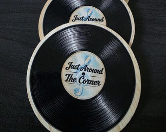 May include: A set of circular coasters designed to resemble black vinyl records with light wood grain borders. Each coaster features a central label with a light blue treble clef graphic and the black text Just Around The Corner.