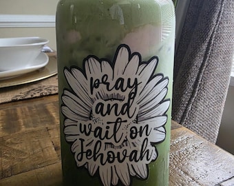 May include: A clear glass tumbler filled with a green beverage, topped with a clear lid and straw. A white and black floral sticker on the glass reads "pray and wait on Jehovah". The drink is on a wooden table.