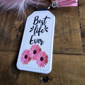 Pode incluir: Chaveiro de couro branco com flores cor-de-rosa e costuras pretas. O chaveiro tem uma al&ccedil;a preta e um tassel rosa. O texto no chaveiro diz "Best life Ever".
