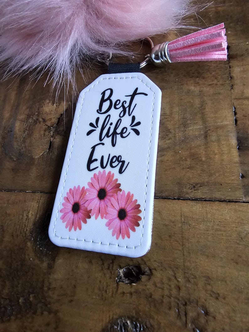 Pode incluir: Chaveiro de couro branco com flores cor-de-rosa e costuras pretas. O chaveiro tem uma al&ccedil;a preta e um tassel rosa. O texto no chaveiro diz "Best life Ever".