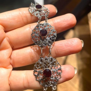 May include: A silver filigree bracelet with three dangling charms. Each charm features a red gemstone surrounded by intricate silver designs.