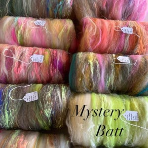 May include: A collection of colorful fiber batts, each a blend of various hues including pink, green, yellow, and blue. The batts are labeled with small tags. The text "Mystery Batt" is displayed in a cursive font.