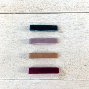 Puede incluir: Cuatro cintas para el pelo de terciopelo en diferentes colores: verde azulado oscuro, morado claro, beige y burdeos. Las cintas para el pelo est&aacute;n dispuestas verticalmente sobre una superficie de madera blanca.