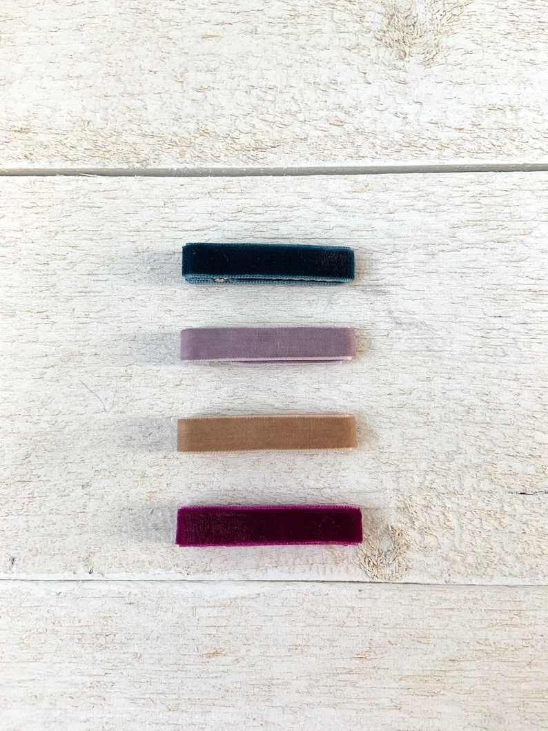 Puede incluir: Cuatro cintas para el pelo de terciopelo en diferentes colores: verde azulado oscuro, morado claro, beige y burdeos. Las cintas para el pelo est&aacute;n dispuestas verticalmente sobre una superficie de madera blanca.