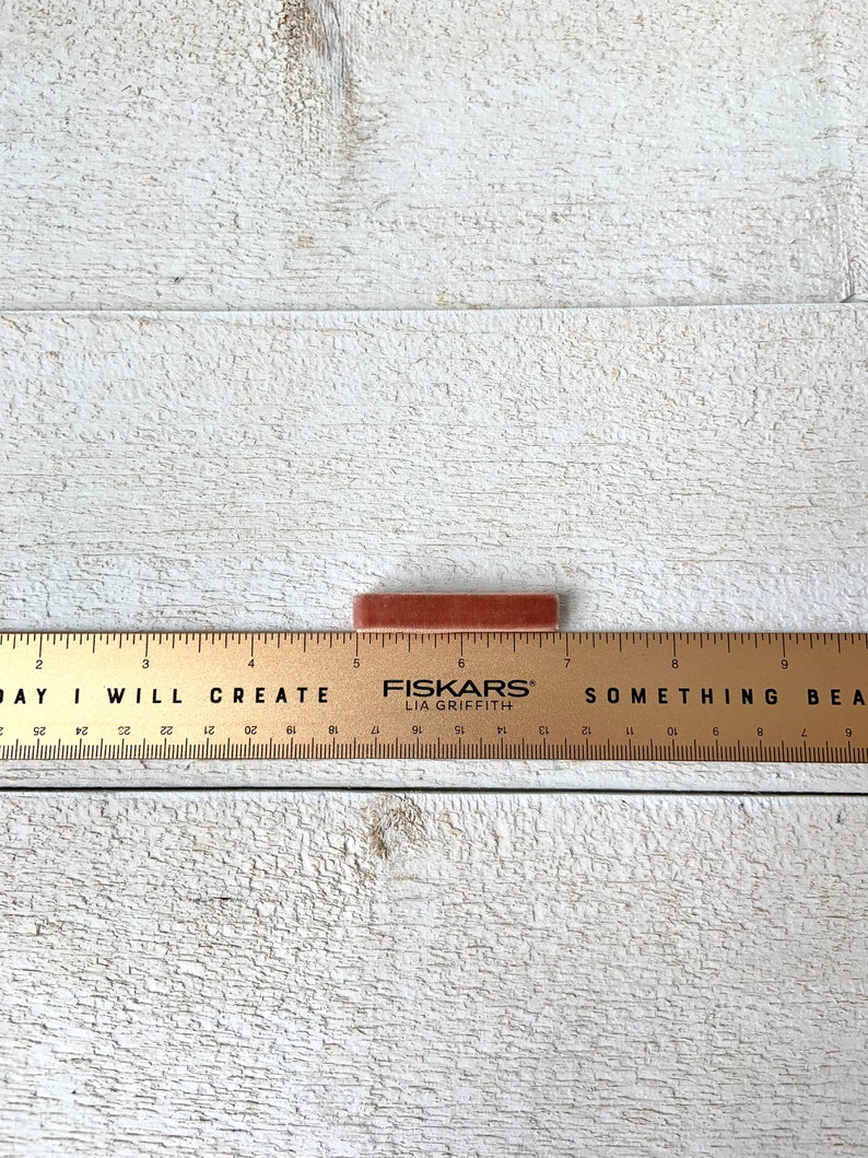 Puede incluir: Un palo de tiza rectangular marr&oacute;n sobre una superficie de madera blanca con una regla que dice "DAY I WILL CREATE" y "FISKARS LIA GRIFFITH" y "SOMETHING BEAUTIFUL".