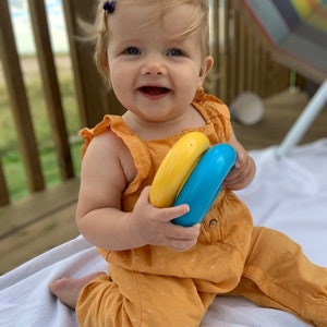 May include: A baby girl in a yellow jumpsuit is sitting on a white blanket and holding two colorful plastic rings. She is smiling and looking at the camera.