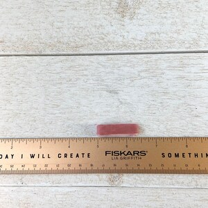 May include: A close-up of a pink eraser with the text "FISKARS&reg; LIA GRIFFITH" printed on it. The eraser is sitting on a wooden surface with a ruler marked with inches.