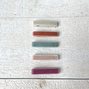 May include: Five velvet hair clips in different colors: beige, terracotta, light blue, blush pink, and burgundy. The clips are arranged vertically on a white wooden surface.