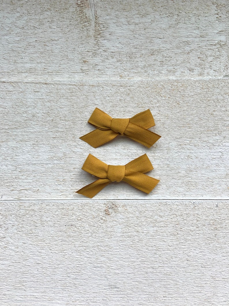 May include: Two mustard yellow fabric hair bows on a white wooden background.