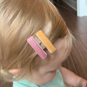 May include: Two hair clips, one pink and one yellow, are in a young person's hair.