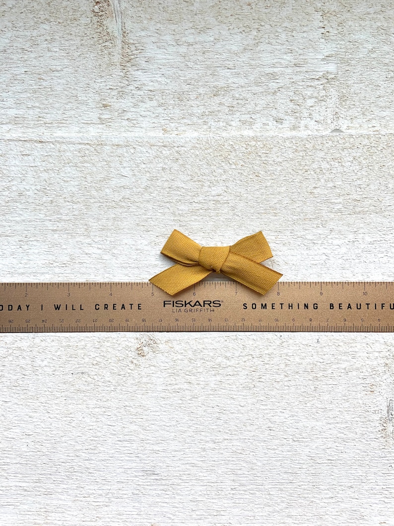 May include: A wooden ruler with the words "TODAY I WILL CREATE SOMETHING BEAUTIFUL" printed on it. A small mustard yellow bow is placed on top of the ruler.